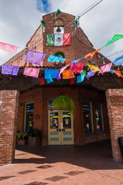A charming brick building decorated with colorful papel picado banners hanging across the front. The Mexican flag is prominently displayed above the entrance, which features double doors and windows on either side. A hanging green plant is centered above the doorway, and the scene is illuminated by natural daylight under a partly cloudy sky.