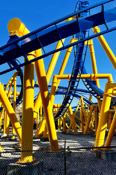 A vibrant roller coaster structure featuring bright yellow support beams and dark blue tracks under a clear blue sky. The coaster twists and turns above a gravel ground, enclosed by a chain-link fence.