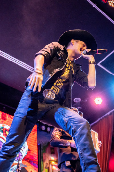 A male singer wearing a large black cowboy hat, black jacket, and dark pants is passionately performing on stage. He holds a microphone close to his mouth and is captured mid-motion with one knee bent and the other leg extended. The stage is lit with colorful lights and there is a vibrant background screen behind him.