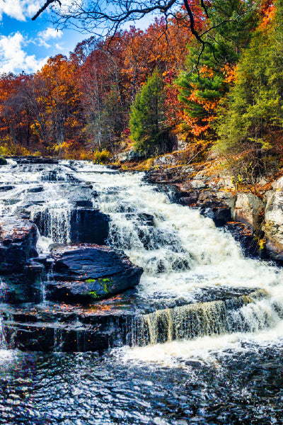 A cascading waterfall flows over dark, rocky ledges surrounded by a forest of autumn foliage in vibrant shades of orange, red, yellow, and green. The sky above is partly cloudy with patches of blue peeking through.
