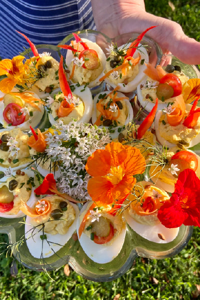 A hand holding a glass platter filled with beautifully decorated deviled eggs. The eggs are topped with colorful garnishes including red chili peppers, small tomato slices, capers, dill, and edible flowers in shades of orange, white, and red, all arranged attractively on the platter. The background shows green grass in natural sunlight.
