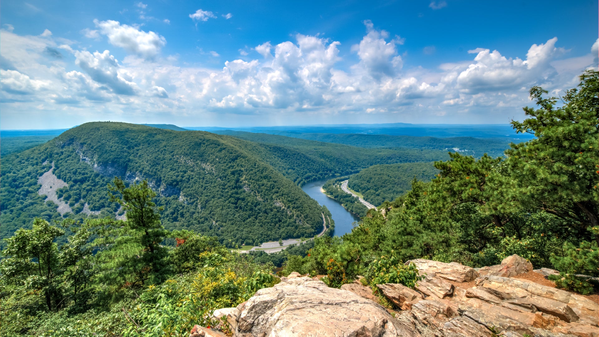 A stunning panoramic view of the Delaware Water Gap, where the Delaware River winds through lush, green mountains. The image is taken from a high vantage point, showcasing rolling hills covered in dense forests under a bright blue sky with fluffy white clouds. In the foreground, rugged rocky terrain is surrounded by vibrant green foliage, adding depth to the breathtaking landscape.