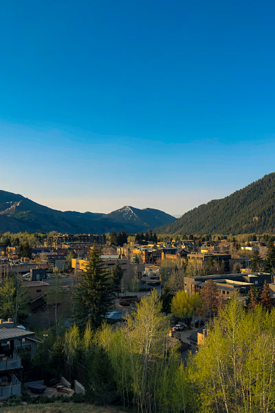 A scenic view of a small mountain town nestled in a valley, surrounded by forested hills and distant snow-capped peaks. The town is bathed in warm, golden light, with a clear blue sky overhead. Trees in the foreground are beginning to show spring foliage, adding a touch of green to the tranquil landscape.
