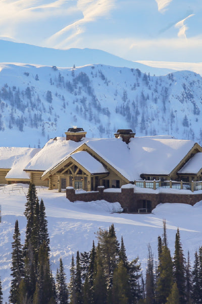 A large mountain cabin covered in a thick layer of snow is nestled among tall evergreen trees. In the background, snow-covered mountain peaks rise against a pale blue sky with soft, wispy clouds.