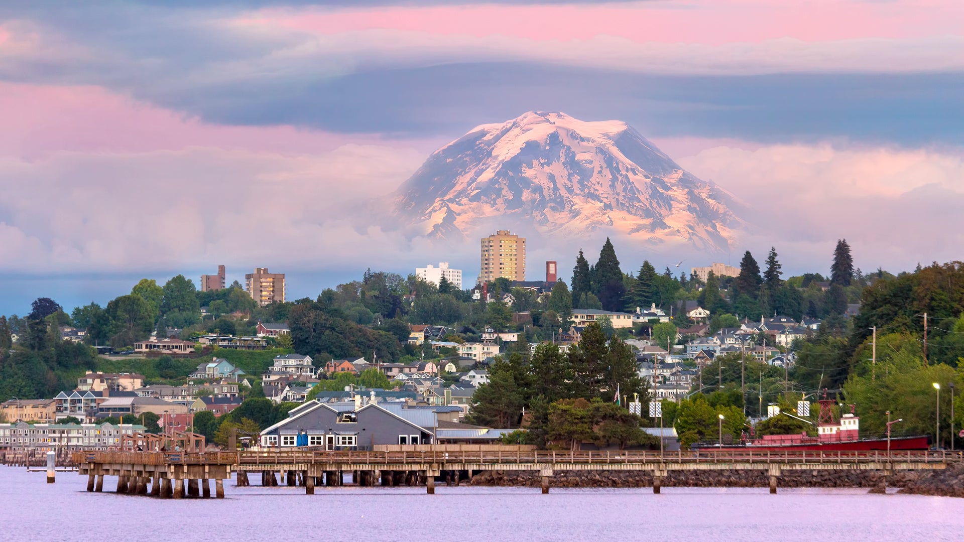 A breathtaking view of Tacoma, Washington, with Mount Rainier towering in the background, partially covered by clouds. The foreground showcases a waterfront with a wooden pier extending into the calm waters, while charming homes and modern buildings are nestled among lush greenery. The sky displays a soft pink and purple hue, adding to the serene and picturesque atmosphere.