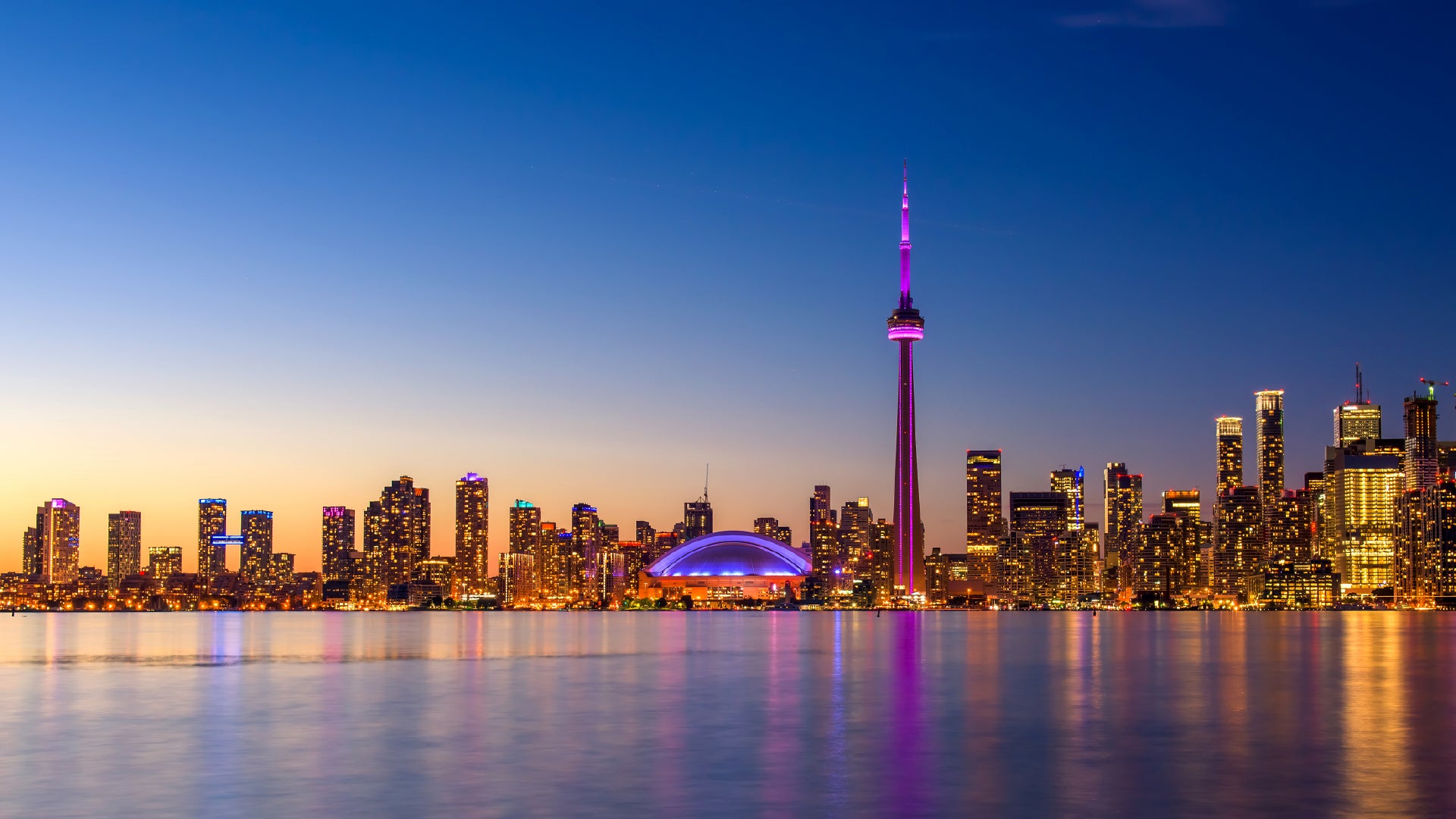 A stunning evening view of Toronto, Canada, featuring the illuminated city skyline with the CN Tower glowing in vibrant purple and the Rogers Centre lit up in blue. The calm waters of Lake Ontario reflect the city lights, creating a beautiful and serene scene against the deep blue and orange hues of the sky.