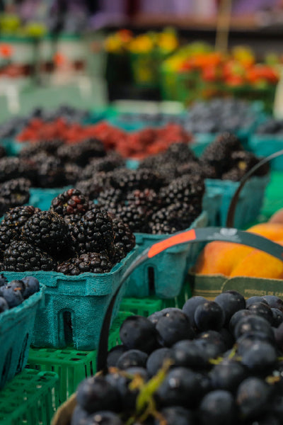Close-up view of fresh blackberries, grapes, and other assorted berries displayed in green cardboard containers at an outdoor farmer's market, with colorful produce blurred in the background.