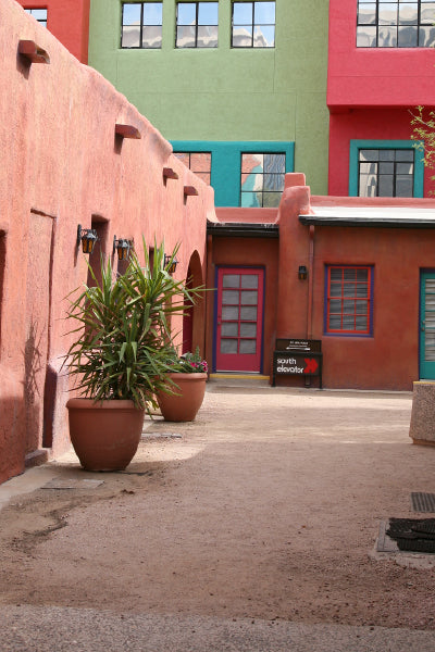A quiet courtyard with adobe-style buildings painted in vibrant red, orange, green, and turquoise, featuring potted plants and a sign indicating the direction of the south elevator.