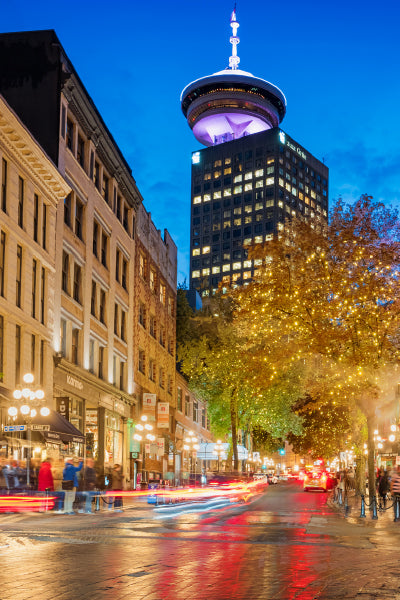 A bustling city street at dusk with historic buildings on the left and trees adorned with twinkling lights on the right. A tall, illuminated tower with a circular observation deck stands prominently in the background against a deep blue sky. Light trails from moving vehicles create dynamic streaks along the wet street.