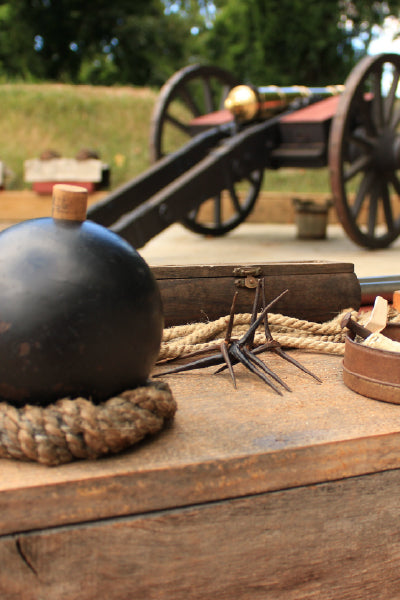 A close-up of an old cannonball with a wooden fuse stopper resting on a coiled rope on a wooden surface. In the background, there is an antique cannon with large wooden wheels and a brass barrel, slightly out of focus. Nearby, there are metal caltrops and other historical items scattered on the wooden surface. The setting appears to be outdoors, possibly at a historical site or reenactment area.