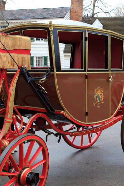 A close-up view of a vintage horse-drawn carriage with elegant red wheels and gold detailing. The carriage body is brown with black trim, featuring a small decorative emblem on the door. The interior is upholstered in red fabric, and the carriage is parked on a paved street with houses in the background.