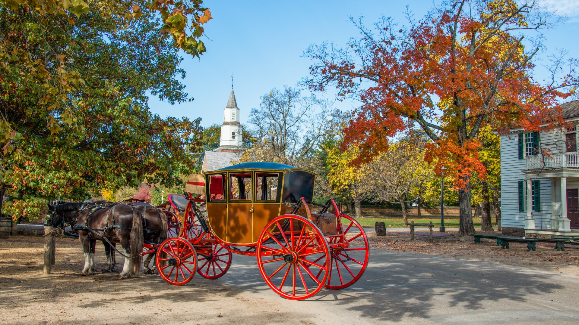 A charming autumn scene in a historic village featuring a vintage horse-drawn carriage with bright red wheels and a brown body. Two dark brown horses stand harnessed to the carriage, waiting near a wooden hitching post. The background showcases a white church with a tall steeple, colonial-style buildings, and trees adorned with vibrant fall foliage in shades of red, orange, and yellow. The warm sunlight filters through the leaves, creating a nostalgic and picturesque atmosphere.