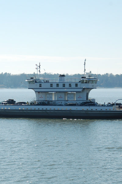 A large, flat ferry boat floating on calm water, designed to transport vehicles and passengers. The ferry has a central two-story cabin with windows and antennas on top. The background shows a distant shoreline with trees under a clear sky.