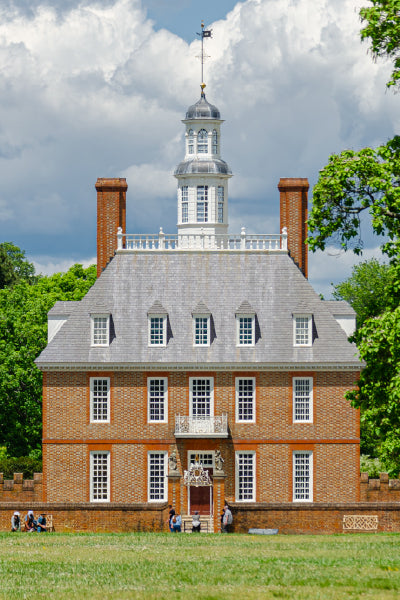 A historic brick mansion with white-framed windows and a tall, multi-tiered cupola on top, set against a partly cloudy sky. The building has a steep gray roof and two brick chimneys. In the foreground, there is a grassy lawn with a few people sitting and walking near the house, and trees surround the property on either side.