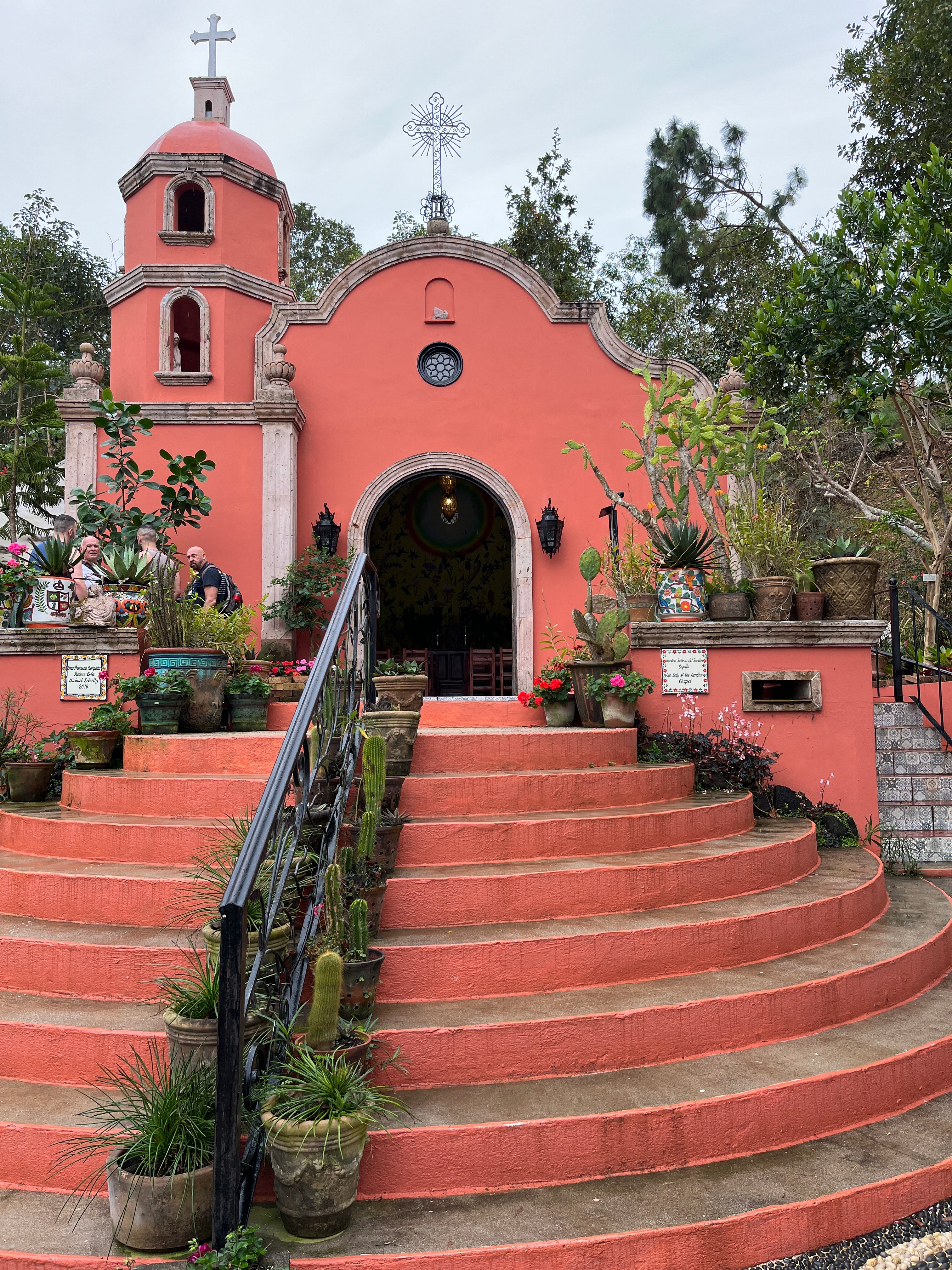 A small, vibrant coral-colored chapel with a bell tower and cross at the top, surrounded by lush greenery. The chapel is accessed by a set of wide, curved steps lined with potted plants and cacti. Decorative pots and flowers adorn the entrance, adding charm and color to the serene setting. The chapel is framed by trees and appears nestled in a peaceful, garden-like environment.