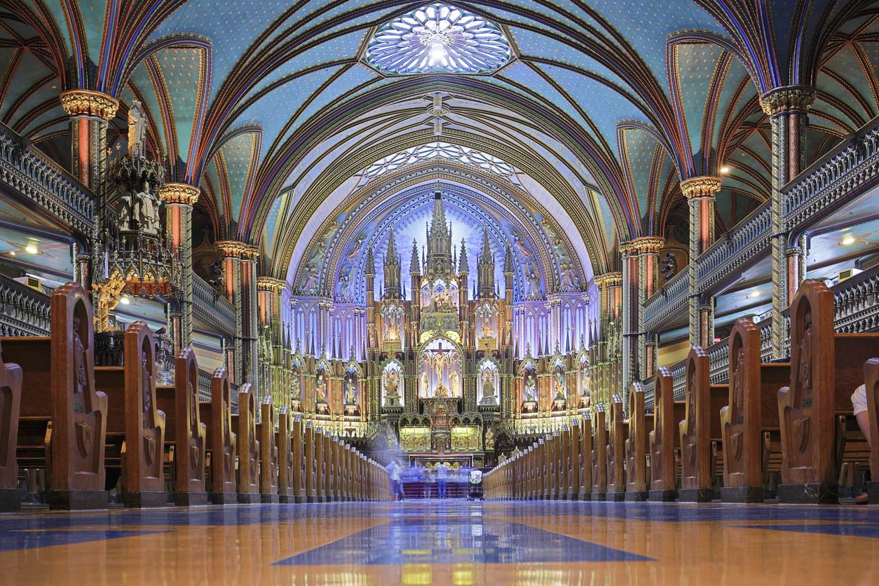 The interior of the Notre-Dame Basilica in Montreal, Quebec, Canada. The basilica is renowned for its stunning Gothic Revival architecture, intricate wood carvings, vibrant stained glass windows, and richly decorated interior with deep blue vaulted ceilings and golden accents. The altar, illuminated dramatically, is a central feature of the church and draws visitors from around the world.